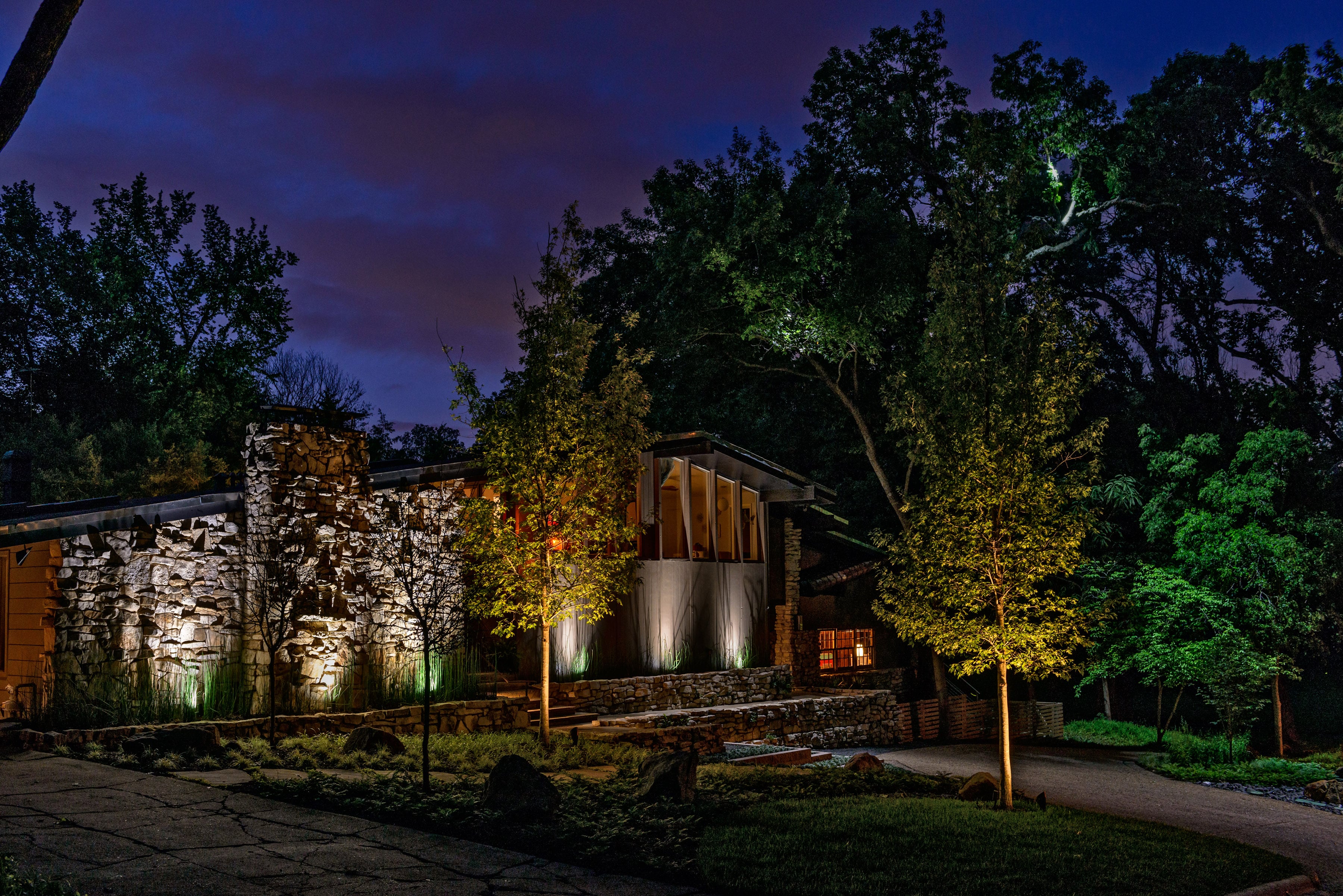 A night-time photo of a home with a custom landscape design with outdoor lighting shining on the trees & home. 